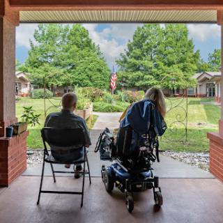 Two individuals enjoying a peaceful moment on the porch, overlooking a green landscape with an American flag in view.