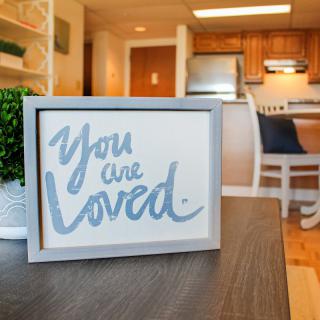 A framed sign in a cozy home setting reads 'You are loved' in front of a kitchen area.