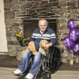 A man in a wheelchair with a dog on his lap, sitting in front of a stone fireplace decorated with balloons.