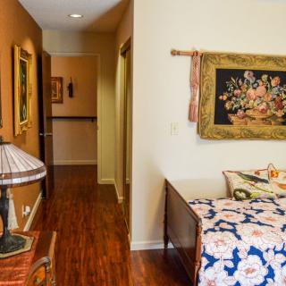 A cozy bedroom with a hallway, featuring wooden flooring, a bed with patterned bedding, decorative artwork on the wall, and a table lamp.