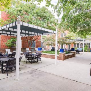 A tranquil outdoor patio with seating areas under a pergola, surrounded by greenery and adjacent to a brick building.