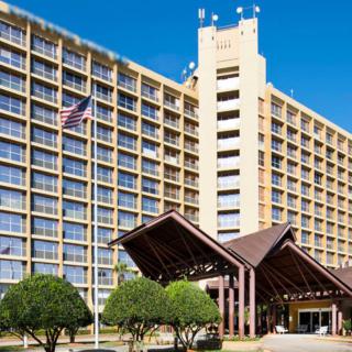A large hotel building with a prominent entrance, surrounded by greenery and featuring an American flag against a clear blue sky.