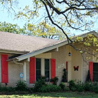 A charming single-story house with red shutters and butterfly decor, surrounded by lush greenery.