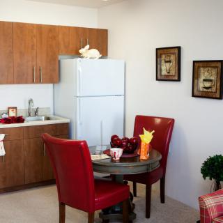 A cozy kitchen and dining area featuring a small table with red chairs, wooden cabinets, and a fridge.