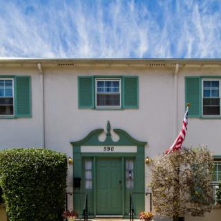 A charming white house with green shutters and a door, displaying an American flag, under a partly cloudy sky.