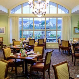A cozy dining room with tables and chairs, featuring a chandelier and large windows allowing natural light.