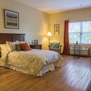 A cozy bedroom with wood flooring, featuring a bed, lamp, and window with curtains.
