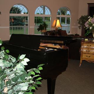 A cozy living room featuring a grand piano, with plants and warm lighting from a lamp next to a set of arched windows.