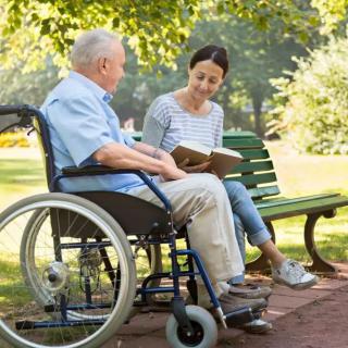 An elderly man in a wheelchair engages in a conversation with a woman reading to him in a park.