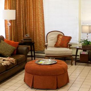 Cozy living room with warm orange and brown tones, featuring a sofa, armchair, and an ottoman in front of large windows.