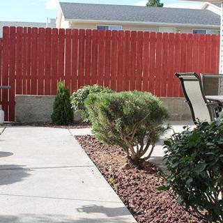 A backyard patio area with a red wooden fence, shrubs, and outdoor furniture.