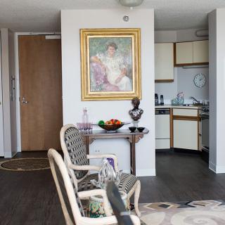 A modern apartment interior featuring a living area with a framed painting, chairs, and a small kitchen in the background.