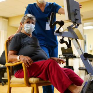 A healthcare professional assists an elderly patient during a rehabilitation session with exercise equipment.