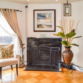 A cozy living room corner featuring a framed artwork above the fireplace, a potted plant, and a comfortable chair by the window.