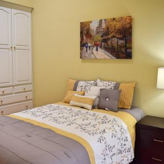 A cozy bedroom with a neatly made bed, decorative pillows, a nightstand with a lamp, and a wall painting above the headboard.