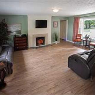A spacious living room with hardwood flooring, featuring a cozy fireplace and a flat-screen TV mounted on the wall. Comfortable seating is arranged around the room, and a small dining area is visible in the background.