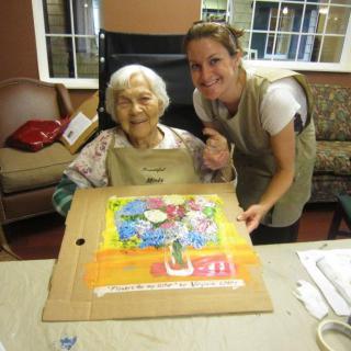 An elderly woman and her caregiver smiling while working on a colorful painting together.
