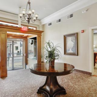 A well-furnished lobby area featuring a round wooden table with a plant, elegant chandelier, and framed artwork on the wall.