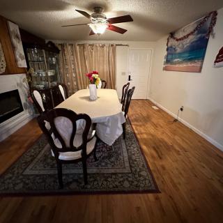 A cozy dining room with a table set for six, featuring a ceiling fan, artwork on the wall, and a decorative rug on wooden flooring.