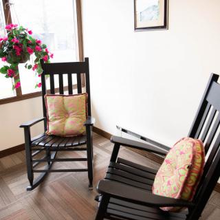A cozy corner with two wooden rocking chairs, adorned with patterned pillows, next to a window with a hanging plant.