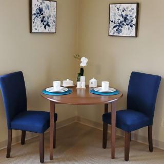 A cozy dining nook with a small round table set for two, featuring blue upholstered chairs and floral wall art.
