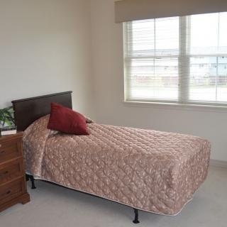 A minimalist bedroom featuring a single bed with a patterned quilt and a pillow, next to a window and a wooden dresser.