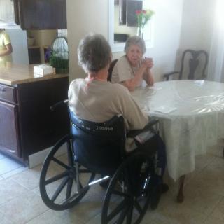 Two elderly women sitting at a table, one in a wheelchair, engaged in conversation.