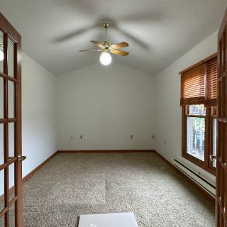 A spacious, empty room with a ceiling fan and large windows, featuring neutral-colored walls and carpet.