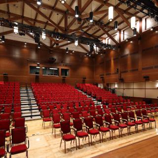 An empty auditorium with red chairs arranged in rows, featuring a wooden interior and stage lighting.