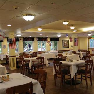 An empty restaurant dining room with tables and chairs neatly arranged, featuring flags and elegant lighting.