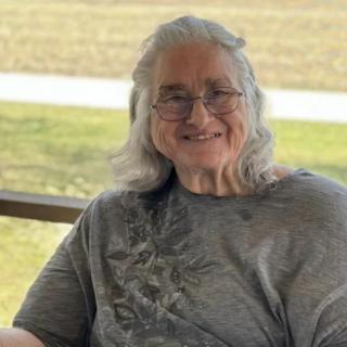 An elderly woman with gray hair and glasses is smiling while seated outdoors.