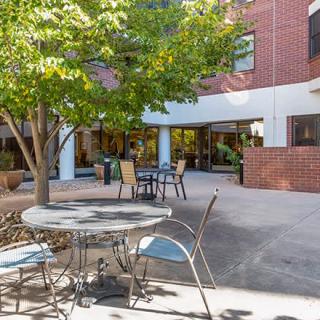 A cozy outdoor patio area with tables and chairs, surrounded by a brick building and shaded by trees.