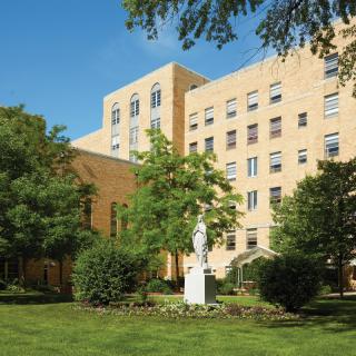 A large building surrounded by trees and a garden with a statue in front on a sunny day.