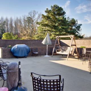 Sunny patio with outdoor furniture, a swing, and a grill surrounded by trees.