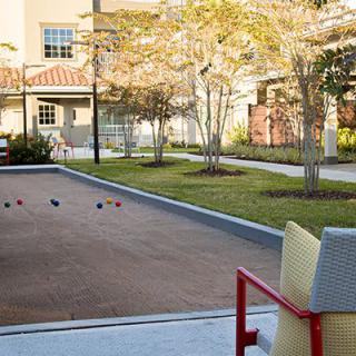An outdoor bocce court with nearby seating, surrounded by a garden and trees in front of a building.