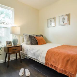 A cozy, minimalist bedroom featuring a neatly made bed with orange accents, a bedside table with a lamp, and a window letting in natural light.