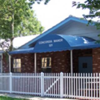 A small church building with a blue awning and a white picket fence surrounded by trees.