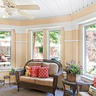 A cozy sunroom with large windows, featuring a rattan sofa, colorful cushions, and decorative plants.