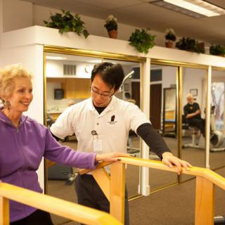 A therapist assists an elderly woman with rehabilitation exercises using parallel bars.