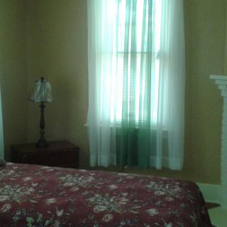 A cozy bedroom featuring a floral bedspread, a window with sheer curtains, and a lamp on the nightstand.
