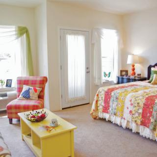 A cozy and colorful bedroom with bright natural light, featuring a bed with a patterned quilt, an armchair, and a small coffee table.