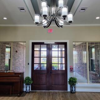 A well-lit interior featuring double wooden doors, a chandelier, and decorative elements including a piano and plants.