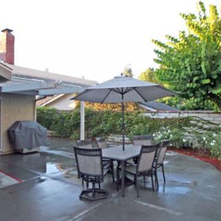 A well-maintained backyard patio with a dining set under an umbrella, surrounded by greenery and featuring a barbecue grill.