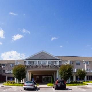 A large building with a symmetrical facade, surrounded by parked cars and trees under a clear blue sky.