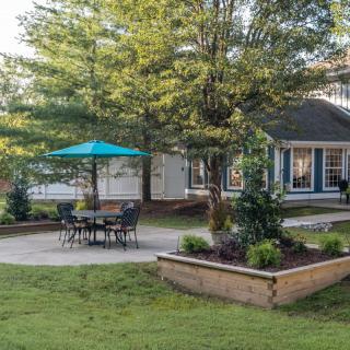 A peaceful backyard garden with a patio set under a blue umbrella, surrounded by lush greenery and a small house in the background.