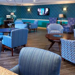 A modern lounge area featuring comfortable seating with blue and patterned chairs, wooden tables, a television on the wall, and ambient lighting from lamps.