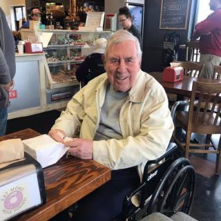 An elderly man in a wheelchair is smiling while seated at a cafe table.