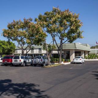 A sunny parking lot with multiple cars and a building surrounded by trees.