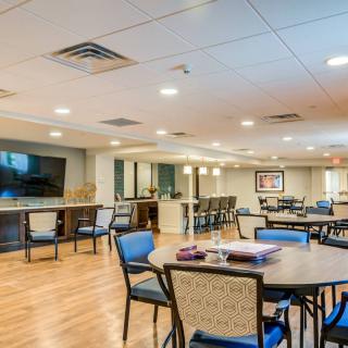 A spacious and well-lit dining area with multiple tables and chairs, featuring a large TV screen on the wall.