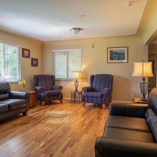 A cozy living room with armchairs and sofas arranged on a wooden floor, illuminated by natural light through the windows.